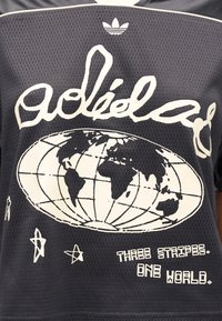 Mustast võrgust t-särk, millel valge maakera graafika ja tekst "Three Stripes, One World." Peal väikse Adidas logo.