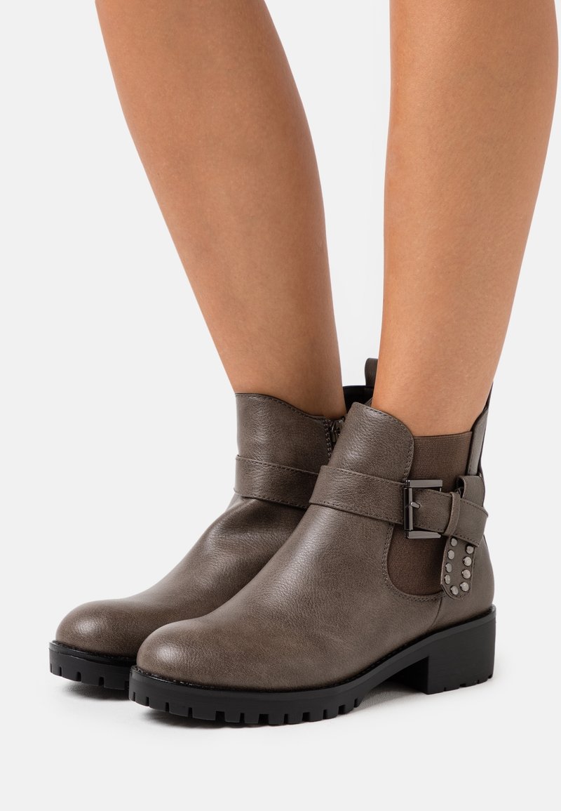 Femme portant des bottines grises avec une boucle latérale et des détails cloutés, debout devant un fond clair uni.