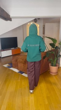Sweatshirt verde com texto "PEGADOR" nas costas, combinada com calças castanhas de pernas largas. Ambiente interior com uma TV, plantas e chão de madeira.