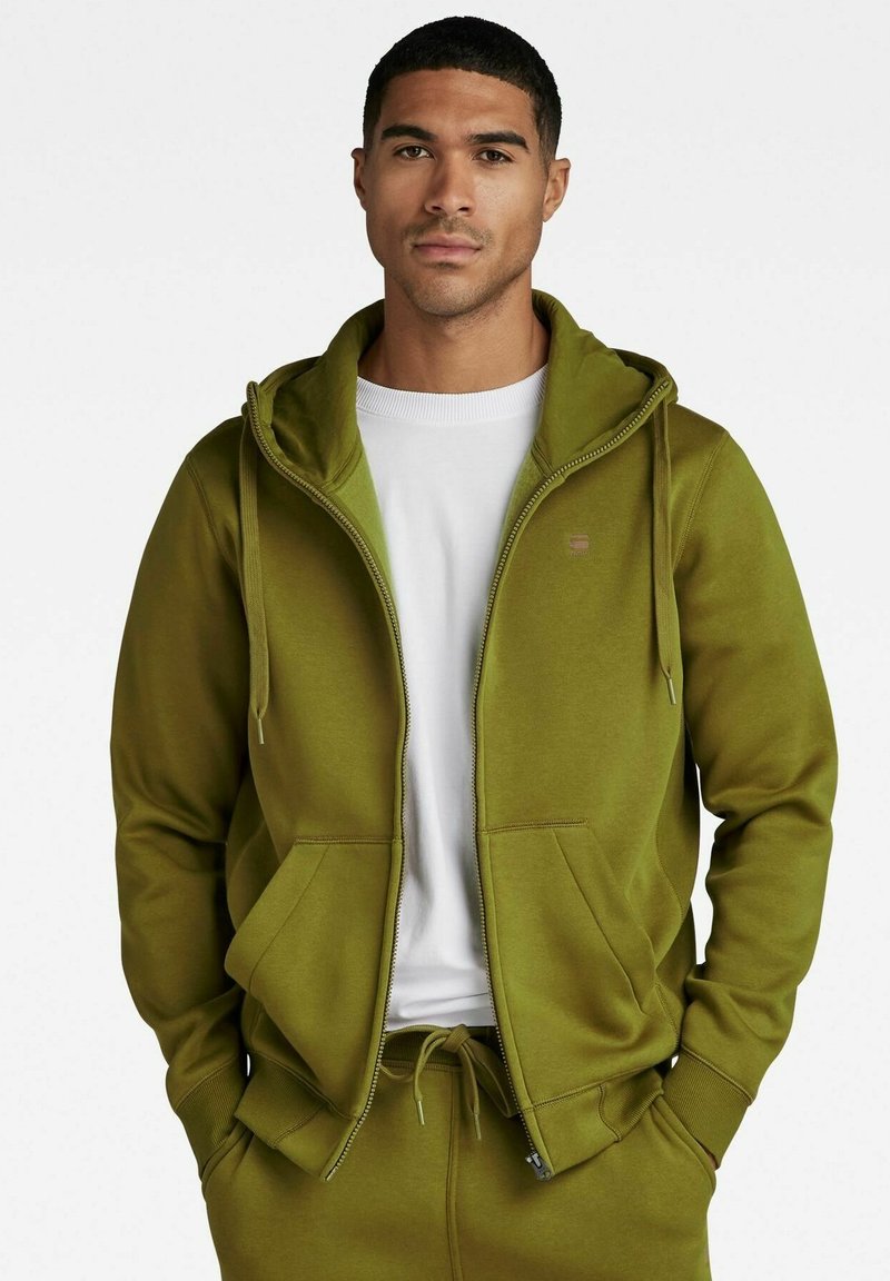 Felpa olive green con zip realizzata in tessuto morbido, dotata di zip frontale, doppie tasche frontali e cappuccio con lacci. Camicia bianca sotto.