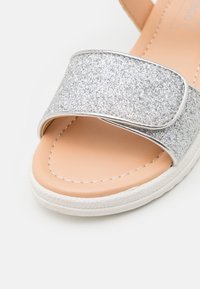 Hopeasa glitterituli, jossa on leveä hihna, vaaleanruskea pohja, valkoinen tekstuuripohja ja koristeellinen ommel reunan ympärillä.