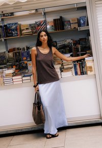 Femme aux cheveux foncés debout tenant un sac fourre-tout marron près du comptoir d'une librairie avec des livres empilés, portant un débardeur marron et une longue jupe blanche.