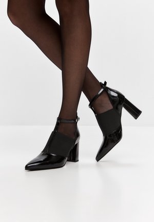 Bottines en cuir verni noir à talons hauts avec côtés élastiques, portées avec des collants noirs transparents sur jambes croisées sur fond blanc uni.