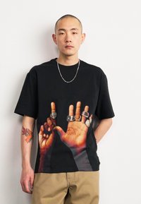 Černé tričko s krátkým rukávem, které má grafický potisk dvou rukou zdobených prsteny. Design zdůrazňuje textury pleti a detaily prstenů.