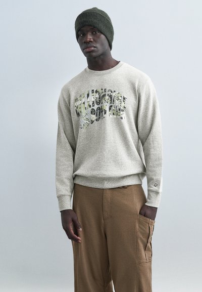 Man wearing olive green knit beanie, gray sweatshirt with graphic text, and brown cargo pants against plain light gray background.