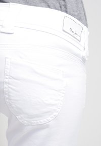 Valkoiset farkut, joissa on istuva muotoilu, pieni takatasku ja huomaamaton ompelukuviot. Brändilogo näkyy vyötärönauhassa.