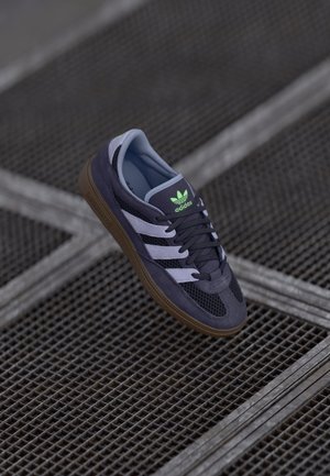 Tmavě modrá teniska značky Adidas se světle modrými pruhy a zeleným logem nad černými tkaničkami, odpočívající na kovovém mřížkovém povrchu.