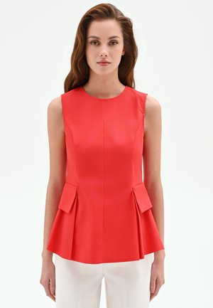 Donna con lunghi capelli castani indossa un top peplum senza maniche, rosso brillante, e pantaloni bianchi, in piedi davanti a uno sfondo bianco uniforme.