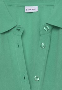 Grüne Bluse mit Kragen aus strukturiertem Stoff. Fünf Knopfverschlüsse, darunter zwei runde, glänzende Knöpfe. Auf dem Etikett steht "LASCANA."