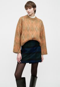 Un pull argyle surdimensionné orange et marron en tissu doux, associé à une mini-jupe à carreaux sombre, des collants noirs et des bottes montantes.