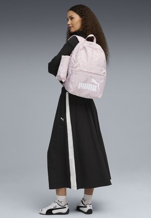 Zaino rosa a fantasia con tasca anteriore, caratterizzato da un logo bianco. Il modello indossa un vestito nero con una striscia bianca e scarpe da ginnastica bianche.