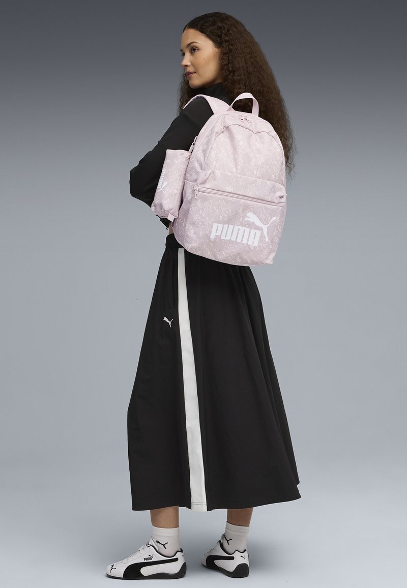 Zaino rosa a fantasia con tasca anteriore, caratterizzato da un logo bianco. Il modello indossa un vestito nero con una striscia bianca e scarpe da ginnastica bianche.