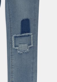 Svetle modré džíny z denimu s opotřebovanými detaily, které mají obdélníkovou nášivku a třepící se okraje. Klasický pětikapsový střih s úzkým pasem.