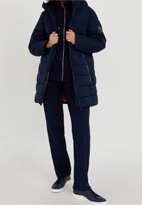 Námořnická puffer bunda s kapucí, prošívaná textura, přední zip a kovové detaily. Nositá přes odpovídající námořnickou mikinu a kalhoty, doplněná teniskami.