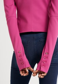 Ruce sepnuté za zády, oblečený těsný jasně růžový blejzr s knoflíkem a detaily manžet v podobě hmyzu, spárovaný s tmavě modrými džínami.