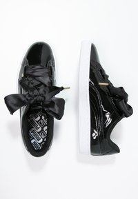 Les baskets en cuir verni noir présentent des lacets satinés noirs surdimensionnés, des accents métalliques dorés et une semelle en caoutchouc blanche. L'intérieur révèle une semelle intérieure à motifs noir et blanc.