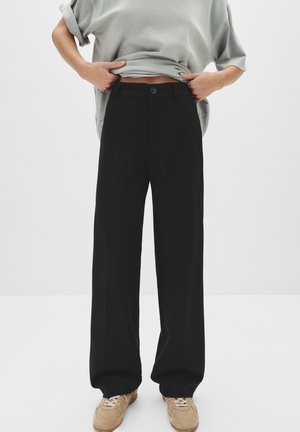 Persona che indossa pantaloni neri larghi e sneakers in suede beige, sollevando una maglietta grigia oversize, su uno sfondo chiaro e semplice.