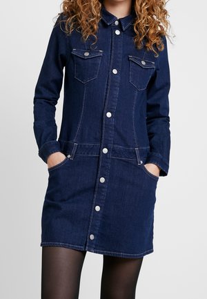Abito di jeans navy con maniche lunghe, bottoni frontali, due tasche sul petto e tasche laterali. Cuciture a contrasto su tutta la lunghezza.