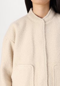 Veste en polaire beige avec un col montant, comportant deux grandes poches avant et une surface texturée et douce. Design simple et fermeture à l'avant.