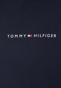 Logo di Tommy Hilfiger con bandiera rossa, bianca e blu centrata su sfondo di tessuto blu navy.