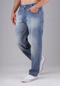 Man wearing faded blue jeans with white sneakers, standing with one hand in pocket and the other relaxed by his side.