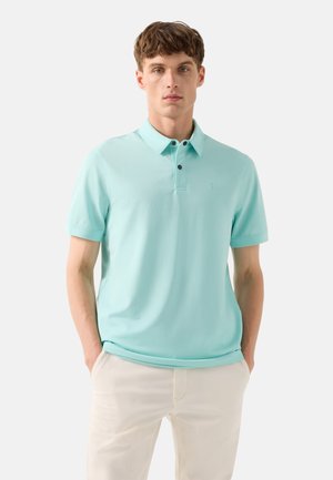 Ung mand iført en lyseblå kortærmet poloshirt og beige bukser, stående med hænderne i lommerne foran en hvid baggrund.