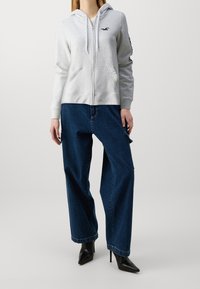 Sudadera gris con cremallera y capucha, con un logo en el pecho, combinada con vaqueros anchos de color azul oscuro y tacones negros de punta afilada.