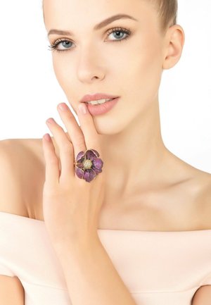Purple flower-shaped ring with intricate detailing and a jeweled center, set against a minimalist background with soft skin tones.