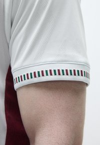 Camisa branca de manga curta com um tecido texturizado e um acabamento listrado em várias cores, incluindo vermelho, verde e branco, ao longo da bainha das mangas.