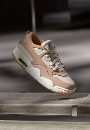 Baskets beiges et blanches avec une semelle épaisse et des lacets, flottant au-dessus d'une surface en béton sous un éclairage intérieur doux.