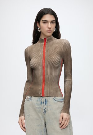 Donna con capelli lunghi e scuri che indossa un top marrone a coste aderente con zip e dettagli rossi e jeans largi chiari.