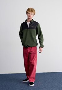 Dark green fleece pullover with a black upper section, zip neckline, paired with loose red pants and black slip-on shoes on a blue carpet.
