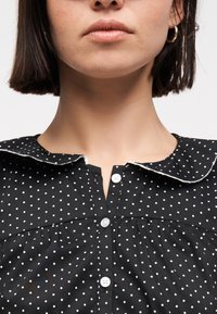 Γυναίκα με κοντά σκοτεινά μαλλιά, που φοράει χρυσούς κρίκους σκουλαρίκια και μαύρο φόρεμα με κουμπιά, λευκές πουά βούλες και στρογγυλή γιακά.