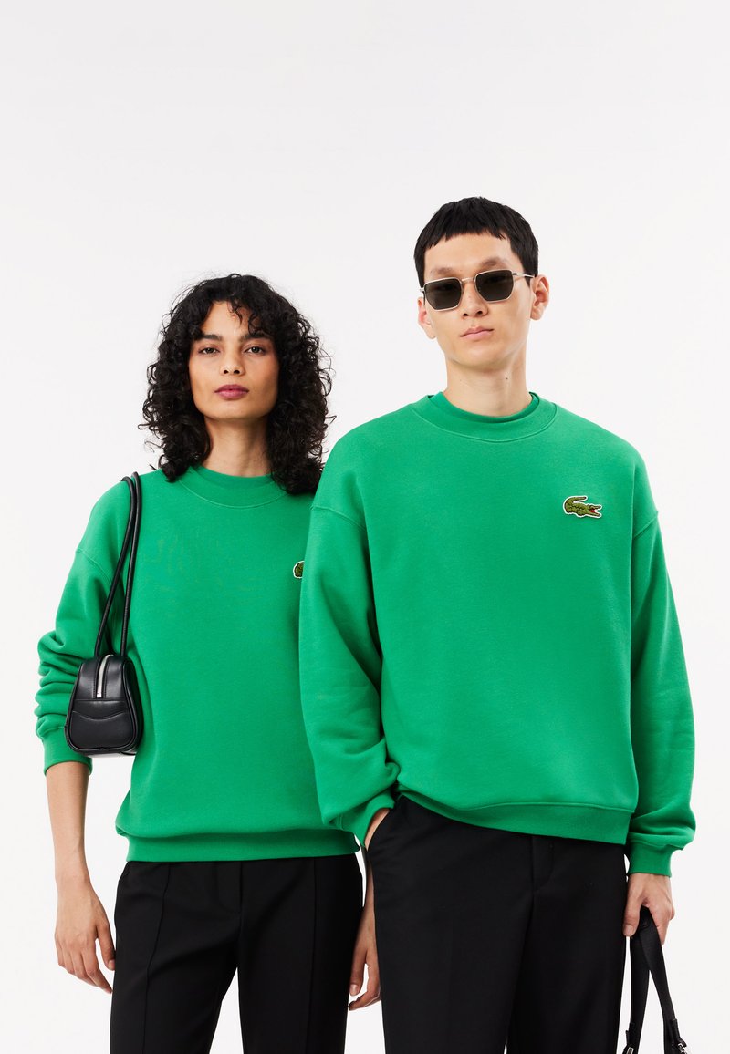 Sudaderas de algodón verdes con cuellos redondos y logotipos bordados, combinadas con pantalones negros. La mujer lleva un bolso negro y tiene el cabello rizado.