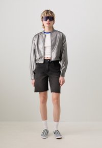 Metallinen hopeinen bomber-takki valkoisen crop-topin päällä, yhdistettynä kulutettuihin mustiin farkkushortseihin ja ruutukuosisisiin liukuvaatteisiin.
