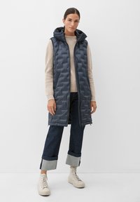 Marineblauer Steppweste mit Kapuze, gestepptem Design, Frontreißverschluss und Seitentaschen, kombiniert mit einem leichten Pullover und hochgekrempelten Jeans.