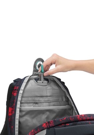 Main tirant une housse de pluie pliable intégrée depuis le compartiment supérieur intérieur d’un sac à dos sombre avec des accents rouges.