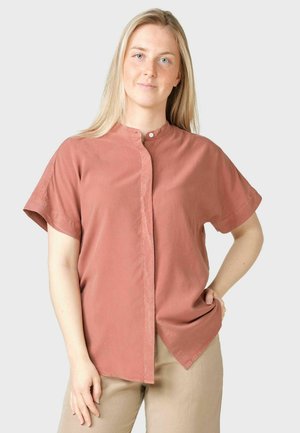 Femme aux longs cheveux blonds portant une chemise à manches courtes couleur rouille et un pantalon beige, debout avec une main sur la hanche.