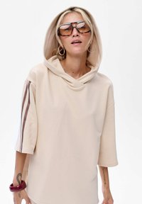 Beżowa oversized bluza z kapturem, krótkimi rękawami i kontrastowymi paskami. Noszona z dużymi okularami przeciwsłonecznymi i bransoletką.