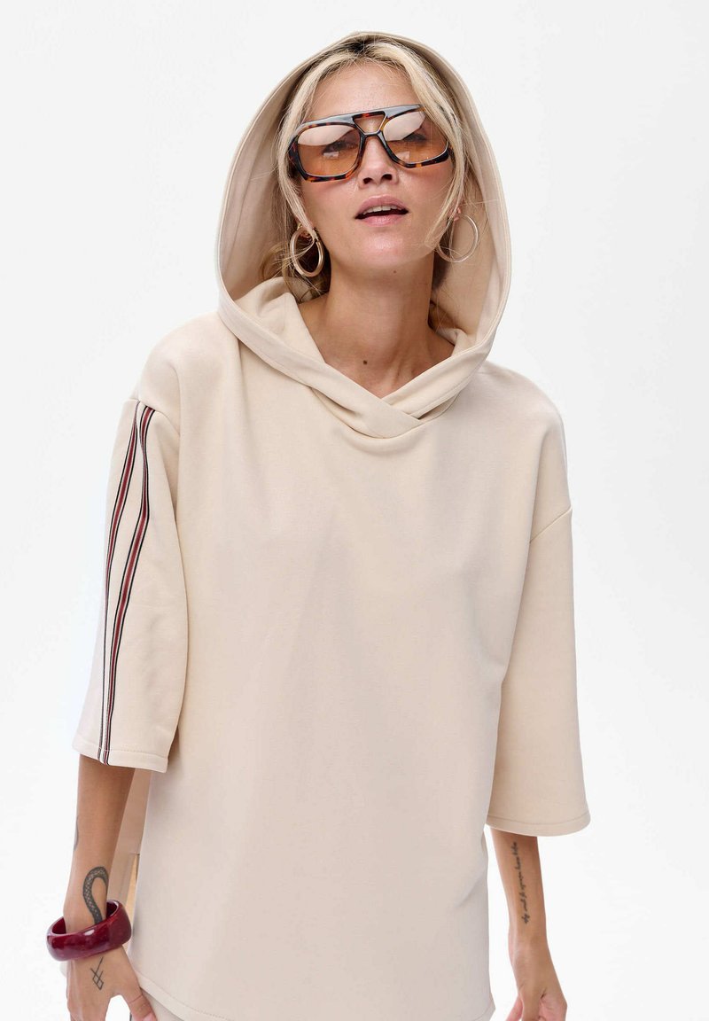 Beżowa oversized bluza z kapturem, krótkimi rękawami i kontrastowymi paskami. Noszona z dużymi okularami przeciwsłonecznymi i bransoletką.