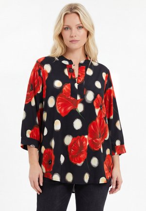 Blonde woman wearing a black blouse with large red poppy flowers and white circular patterns, standing against a white background.