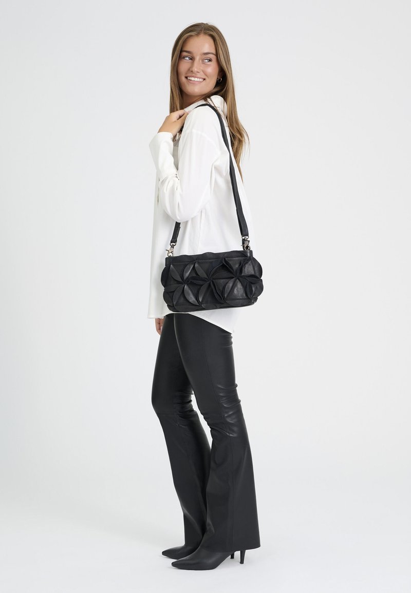 Femme portant un chemisier blanc et un pantalon en cuir noir, portant un sac à épaule noir texturé, regardant par-dessus son épaule et souriant.