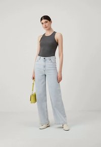 Noor naine tumepruunide juustega hallis ilma varrukateta t-särgis, heledates laiadest pükstest, valgetes kingades, hoiab väikest kollast käekotti, seistes ühevärvilisel taustal.