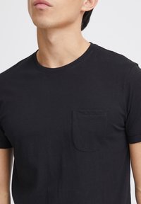 T-shirt en coton noir avec un col rond et des manches courtes. Présente une poche sur la poitrine gauche avec un ourlet droit et une texture douce.