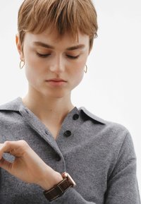 Suéter de lana gris de punto con collar y botones negros, que lleva una persona que está revisando un reloj marrón con cara metálica en su muñeca.