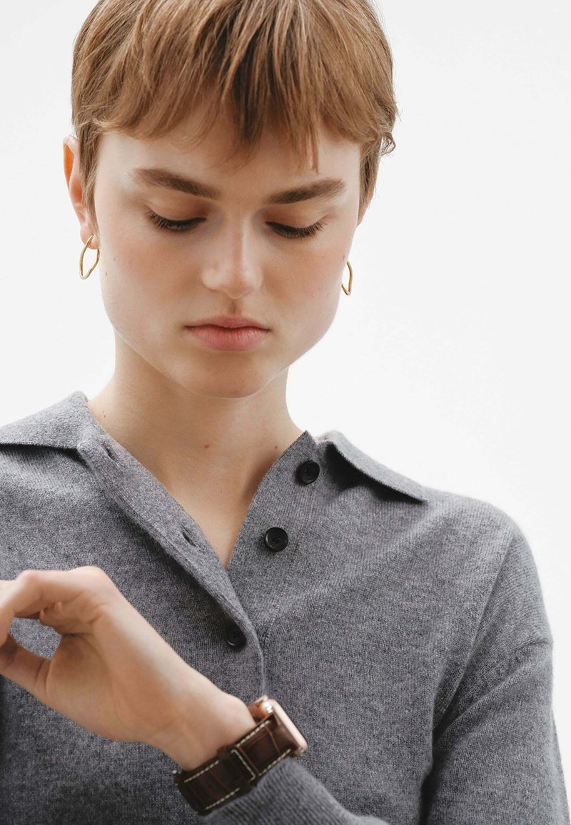 Suéter de lana gris de punto con collar y botones negros, que lleva una persona que está revisando un reloj marrón con cara metálica en su muñeca.