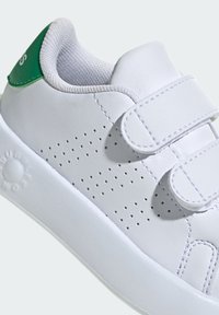 Zapatilla blanca con parte superior de cuero perforado, dos correas de Velcro, cuello acolchado y acento verde en el talón. Suela de goma sólida.