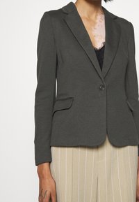 Ciemnoszara marynarka z klapami z wycięciem, zapinana na jeden guzik, z dwoma bocznymi kieszeniami i fakturowaną tkaniną. W zestawie z beżowymi spodniami w prążki.