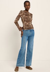 Frau in einem langärmeligen Oberteil mit Leopardenmuster und passenden Schuhen, kombiniert mit hochgeschnittenen, weiten blauen Jeans, vor einem schlichten Hintergrund stehend.