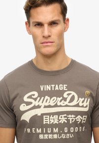 Camiseta de algodón gris con un gráfico de texto blanco que dice "Vintage SuperDry" con un acento amarillo y textura desgastada. Cuello redondo.